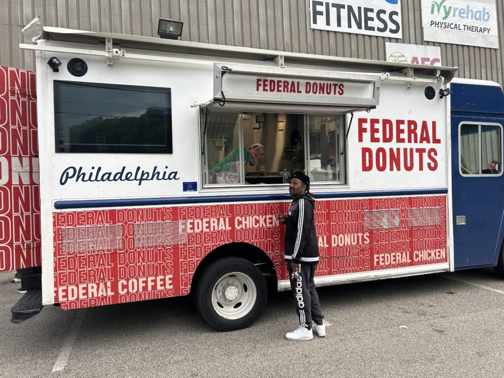 A large red, white, and dark blue food truck that says "Federal Donuts" in red. There is someone inside the truck smiling and someone at the window ordering food.