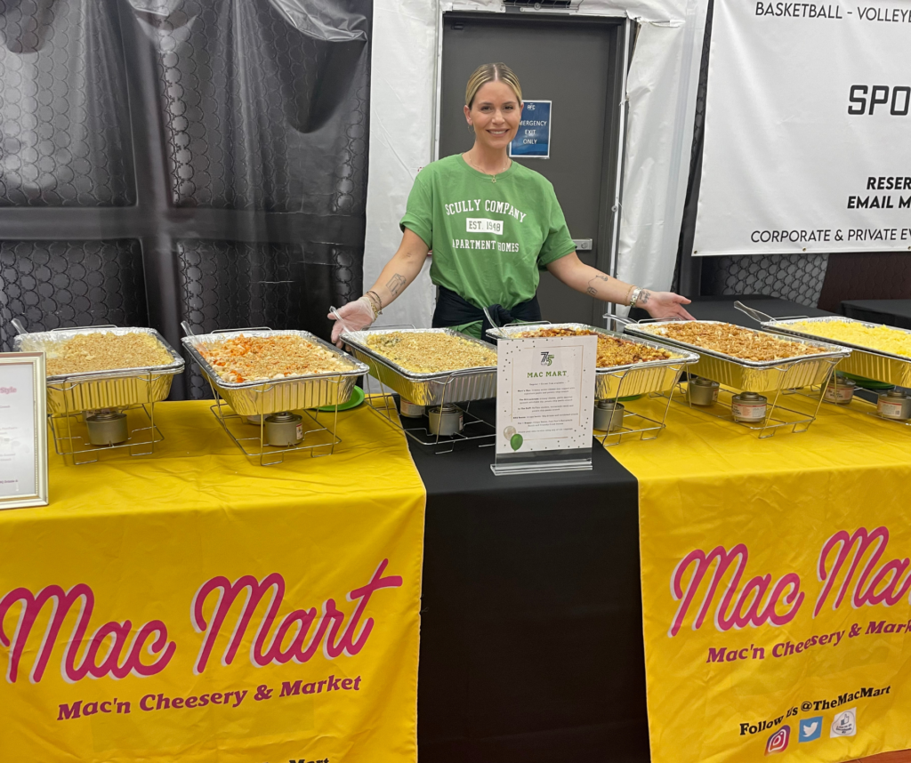 A woman in a green Scully shirt smiling and standing in front of a table full of catering trays with mac and cheese.