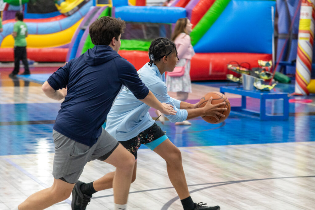 Two young teenagers playing basketball in front of several bouncy houses