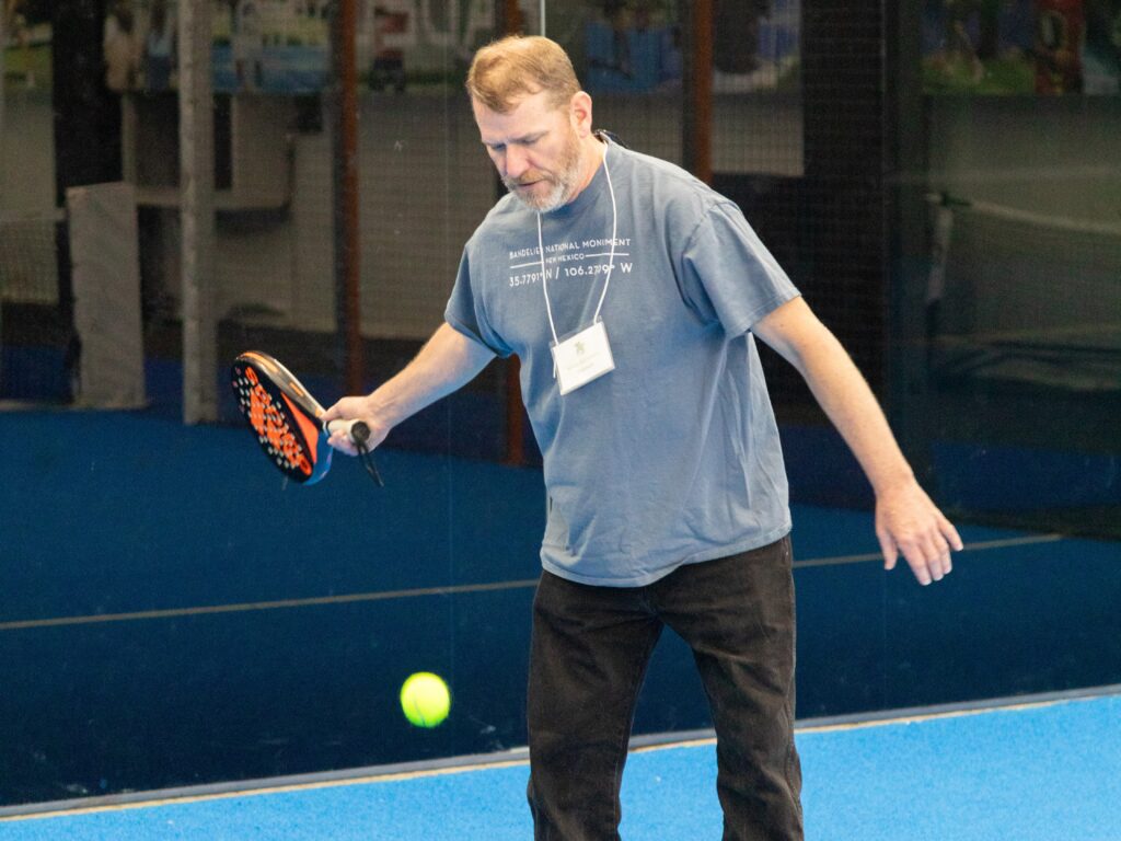 A man with a padel paddle in his hand about to hit a tennis ball on a blue court surrounded by glass