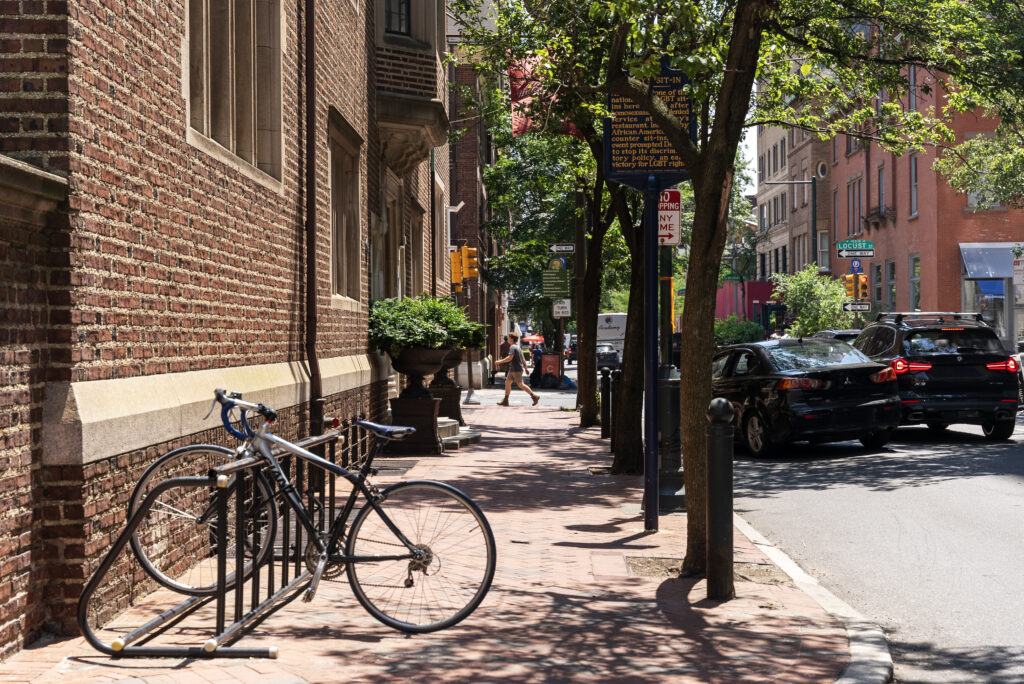 A sunny Philadelphia sidewalk with a parked bike and people walking next to brick buildings