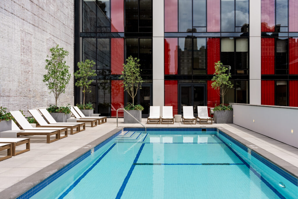 Blue swimming pool with white lounge chairs surrounding it. Next to a white brick wall and a glass building